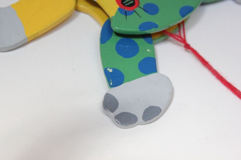 Puede incluir: Un t&iacute;tere de madera de un gato con pintura verde, azul y amarilla. El gato tiene una cuerda roja atada a su espalda.