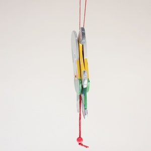 Puede incluir: Un juguete de madera con una cuerda roja y una cuenta roja colgando de &eacute;l. El juguete est&aacute; pintado en tonos de azul, verde, amarillo y blanco. El juguete tiene la forma de un p&aacute;jaro con las alas extendidas.