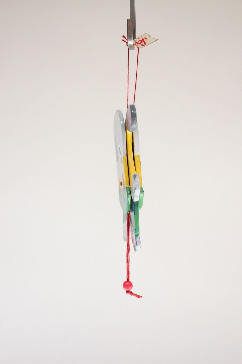 Puede incluir: Un juguete de madera con una cuerda roja y una cuenta roja colgando de &eacute;l. El juguete est&aacute; pintado en tonos de azul, verde, amarillo y blanco. El juguete tiene la forma de un p&aacute;jaro con las alas extendidas.