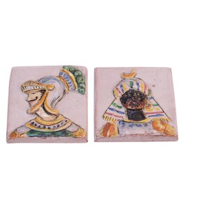 May include: Two ceramic tiles with raised images of people. The tile on the left features a person wearing a helmet and armor, while the tile on the right features a person wearing a colorful headdress.