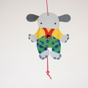 Puede incluir: Un t&iacute;tere de madera de un perro colgado de un gancho. El perro est&aacute; pintado de blanco, amarillo, verde, azul y rojo. Tiene la nariz roja y los ojos negros. El perro lleva una camisa amarilla y pantalones verdes con lunares azules.