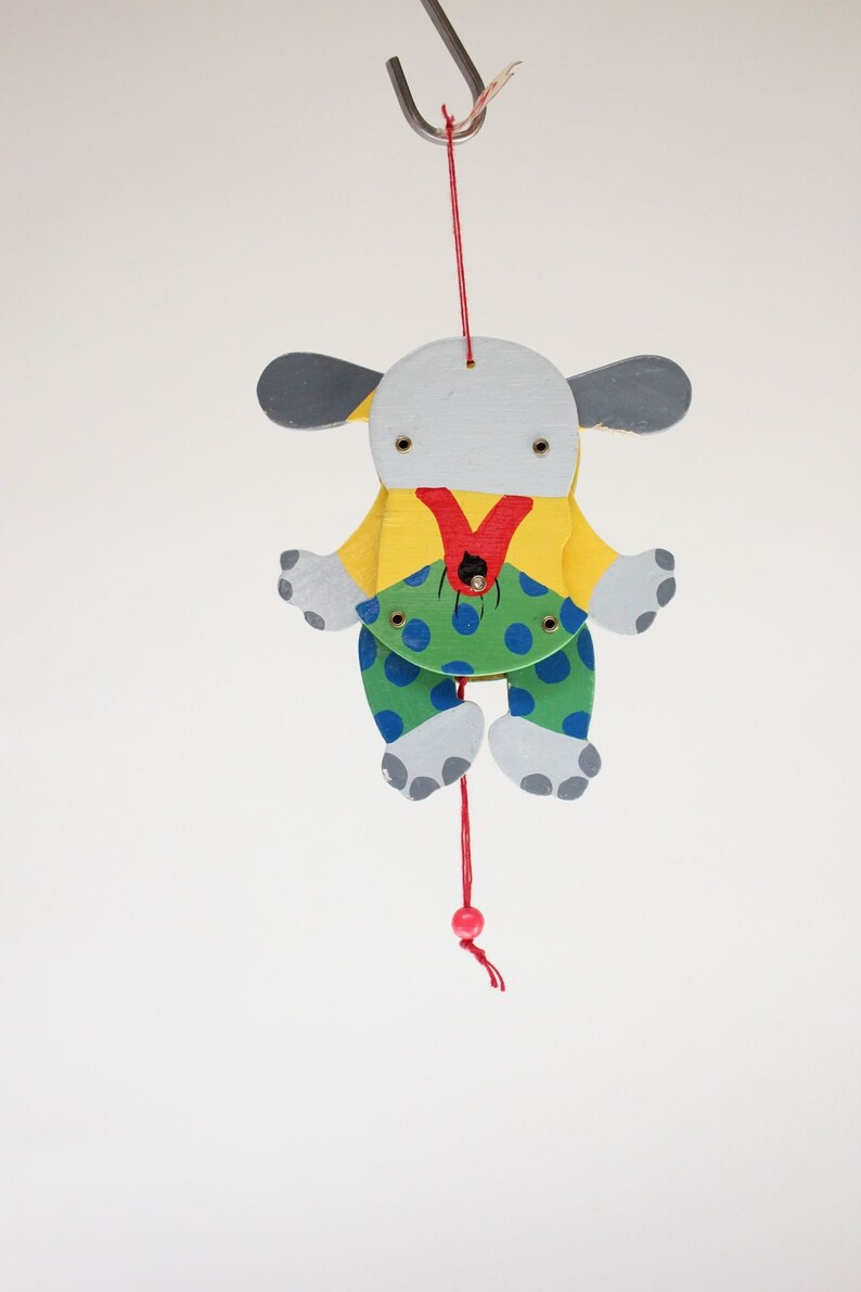 Puede incluir: Un t&iacute;tere de madera de un perro colgado de un gancho. El perro est&aacute; pintado de blanco, amarillo, verde, azul y rojo. Tiene la nariz roja y los ojos negros. El perro lleva una camisa amarilla y pantalones verdes con lunares azules.