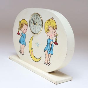 May include: A vintage, oval-shaped clock with a cream-colored finish. The clock face is round with a gold-tone bezel. The clock is decorated with cartoon figures of children holding candles and a yellow crescent moon. The clock sits on a rectangular base.