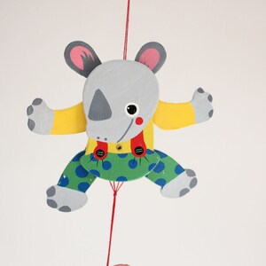 Puede incluir: Un t&iacute;tere de madera gris, amarillo, verde y rojo con forma de rinoceronte con una cuerda atada a su cabeza. El t&iacute;tere lleva una camisa amarilla, tirantes rojos y pantalones verdes con lunares azules.