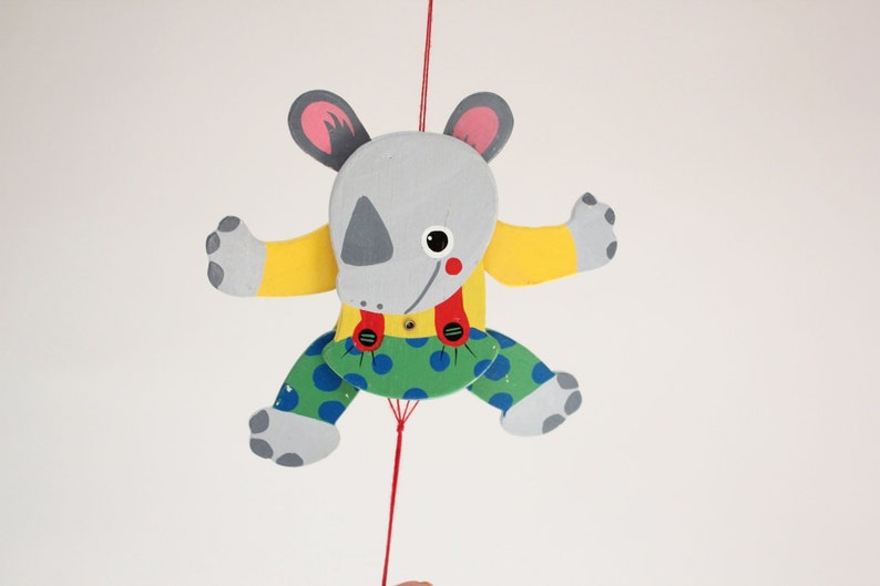 Puede incluir: Un t&iacute;tere de madera gris, amarillo, verde y rojo con forma de rinoceronte con una cuerda atada a su cabeza. El t&iacute;tere lleva una camisa amarilla, tirantes rojos y pantalones verdes con lunares azules.