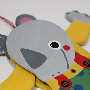 Puede incluir: Un juguete de madera en forma de animal con cabeza gris, cuerpo amarillo y ropa con estampado verde y azul. El juguete tiene boca roja, ojos negros y un collar rojo y amarillo.