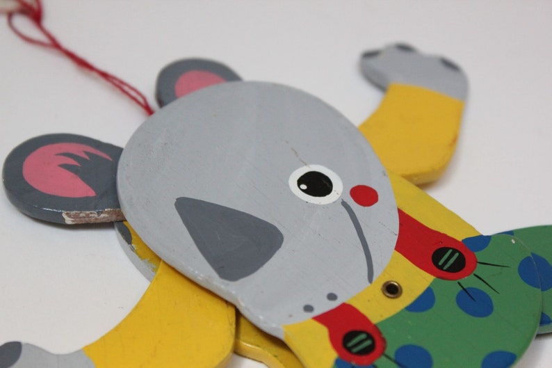 Puede incluir: Un juguete de madera en forma de animal con cabeza gris, cuerpo amarillo y ropa con estampado verde y azul. El juguete tiene boca roja, ojos negros y un collar rojo y amarillo.