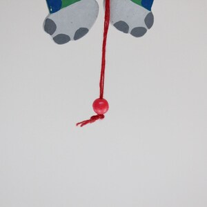 Puede incluir: Una cuenta de madera roja cuelga de una cuerda roja unida a un par de pies de madera pintados de gris y verde.