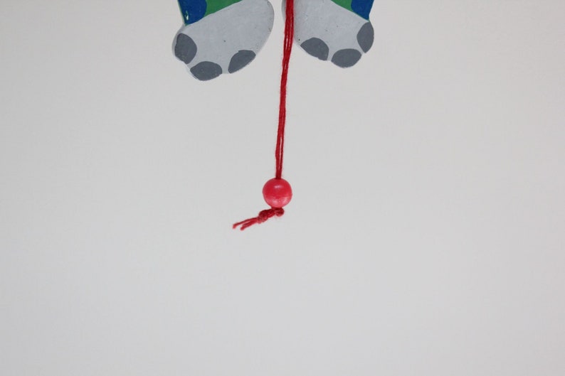 Puede incluir: Una cuenta de madera roja cuelga de una cuerda roja unida a un par de pies de madera pintados de gris y verde.