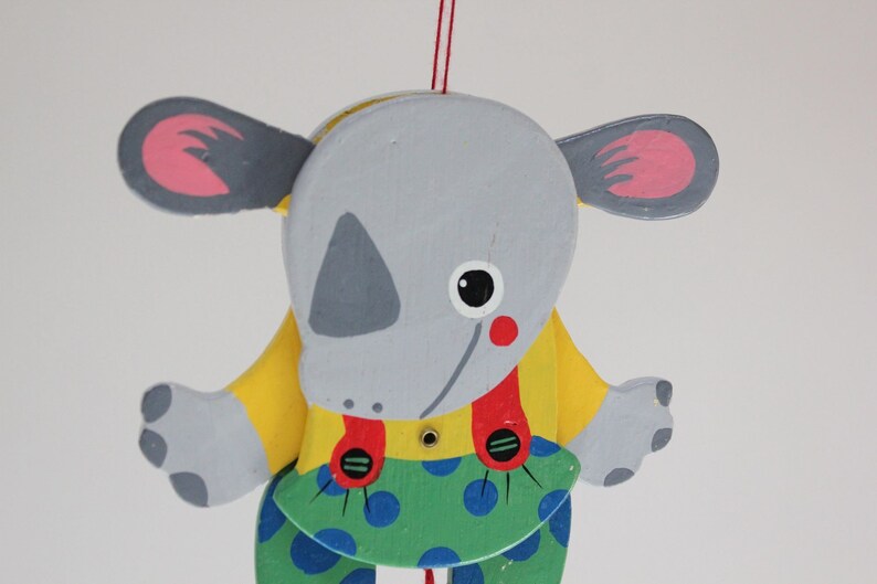 Puede incluir: Un t&iacute;tere de madera gris y amarillo de un animal de dibujos animados con orejas grandes, una nariz roja y pantalones de lunares azules y verdes. El t&iacute;tere est&aacute; colgado de una cuerda.