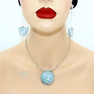 Könnte beinhalten: Ein silbernes Halsketten- und Ohrring-Set. Die Halskette hat einen runden, hellblauen Anhänger mit Sternmuster. Die Ohrringe haben ein ähnliches hellblaues Design in Halbmondform, das an silbernen Drähten hängt. Der Schmuck wird auf einer Schaufensterpuppe präsentiert.