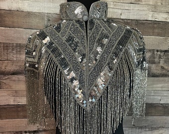 May include: A silver sequined and beaded shawl with fringe. The shawl has a geometric pattern and a high collar.