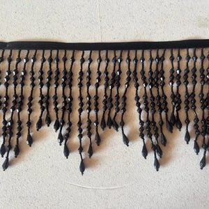 Könnte beinhalten: Schwarze Perlenfransen. Die Verzierung hat eine schwarze Stoffbasis mit Strängen aus schwarzen Perlen, die herunterhängen. Die Perlen sind eine Mischung aus Formen, darunter facettierte und tropfenförmige Designs. Diese Verzierung eignet sich für verschiedene Bastel- und Designprojekte.