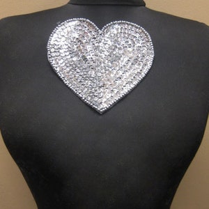 May include: A silver heart-shaped sequin patch. The patch is on a black background.
