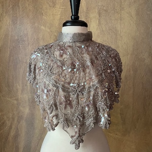 May include: A silver-gray beaded capelet with a high collar, displayed on a mannequin. The capelet features intricate floral and leaf patterns, embellished with sequins. The delicate design suggests formal wear or vintage style.