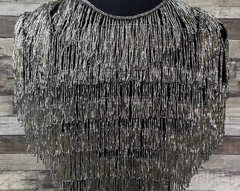 May include: A silver beaded fringe collar necklace. The necklace is made of small, silver beads that are strung together to create a fringe effect. The necklace is worn on a black mannequin.