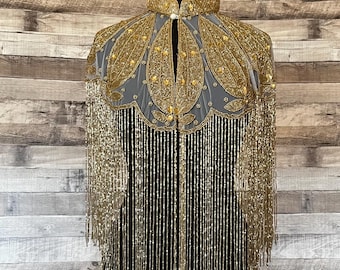 May include: A gold beaded cape with a fringe trim. The cape has a high collar and a beaded design.