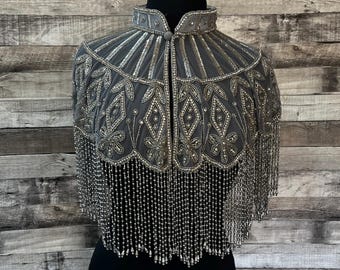 May include: A grey beaded cape with a high collar and button fastening. The cape features detailed silver beadwork in floral and geometric designs. Long strands of silver beads form a fringe along the bottom edge.
