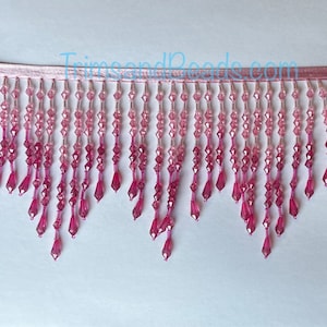 May include: Pink beaded fringe trim with a gradient of colour from light to dark pink. The fringe is attached to a thin pink ribbon.