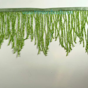 May include: Green beaded fringe trim with a satin ribbon backing. The fringe is made of small, light green beads, creating a textured, decorative element. The trim is suitable for embellishing clothing, accessories, or home decor projects.