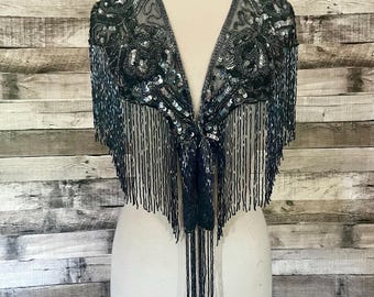 May include: A dark teal beaded shawl with fringe. The shawl is displayed on a mannequin. The top of the shawl is heavily embellished with sequins and beads. The fringe hangs down from the bottom of the shawl.
