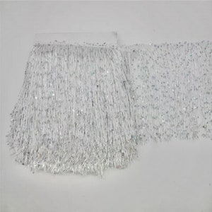May include: White fringe trim with silver sequins. The trim features long, thin strands of white material, interspersed with small, round silver sequins. The fringe is attached to a white backing.