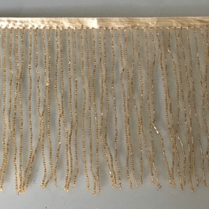 May include: A close-up of a beige beaded fringe trim. The fringe is made of small, square beads that are strung together on a thin thread. The fringe is about 1 inch wide and has a smooth, silky texture.