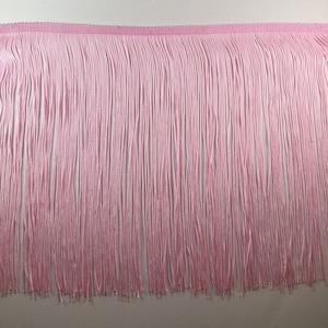 May include: A length of pale pink fringe trim. The trim has a woven header and long, vertical strands of soft pink thread. The fringe is densely packed, creating a full and textured appearance. Suitable for adding a decorative element to clothing or home decor projects.