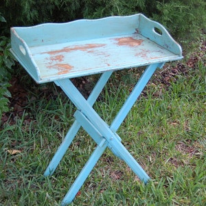 Op de afbeelding: Een lichtblauwe houten klaptafel met een dienblad. De tafel is verweerd en heeft een rustieke uitstraling. Hij staat op een bed van groen gras.