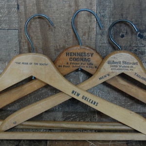 Puede incluir: Tres perchas de madera vintage con ganchos de metal. La percha superior est&aacute; etiquetada con "The Roosevelt, The Pride of the South". La percha del medio est&aacute; etiquetada con "Hennessy Cognac, A Friend For Life, 84 Proof Schieffelin & Co., N.Y.". La percha inferior est&aacute; etiquetada con "Robert Stewart, 1600 Walnut St., Philadelphia, Pa., Men's Tailors".