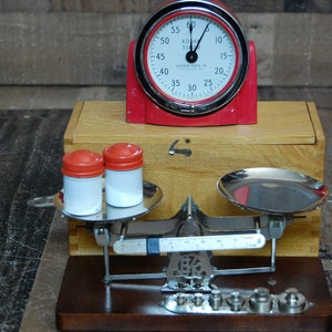 May include: A vintage red kitchen timer with a white face and black numbers sits on top of a wooden box. A silver balance scale with two round metal plates sits on a wooden base. Two white containers with red lids are on the scale's left plate.