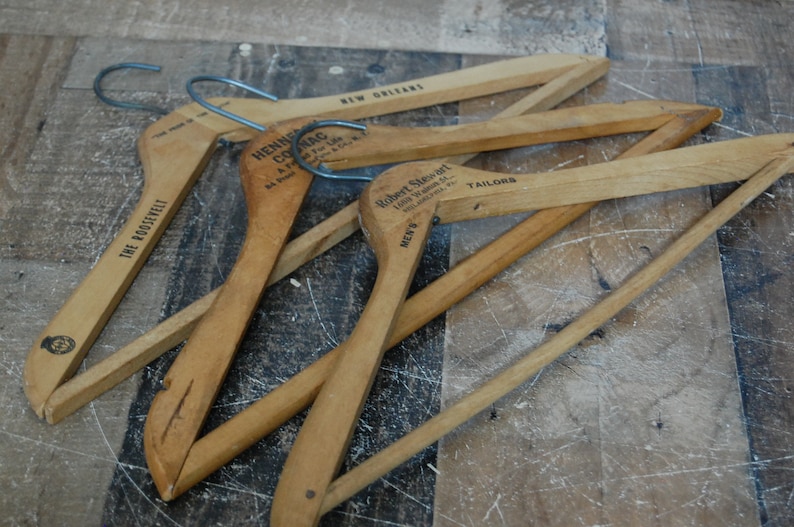 Puede incluir: Cuatro perchas de madera con diferentes etiquetas. Las perchas son de madera natural y tienen un dise&ntilde;o sencillo. Las etiquetas incluyen "The Roosevelt", "Hennig Clothing Co.", "Robert Sievert Tailors" y "New Orleans".