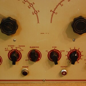 May include: A close-up of a vintage electronic device with multiple knobs and dials. The device has a cream-colored face with red markings and black knobs. The text on the face reads "Stand-By Hi", "Range Lo", "Blanking", "X100", "X10K", "Amplitude", and "Mark".