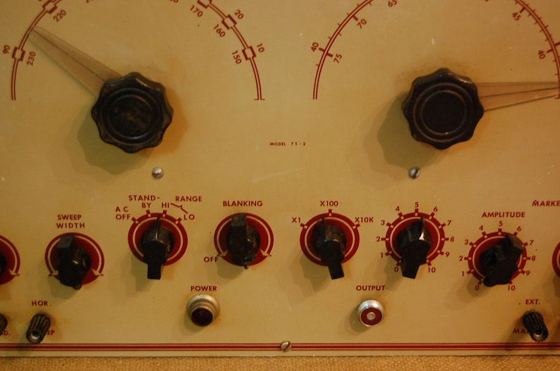 May include: A close-up of a vintage electronic device with multiple knobs and dials. The device has a cream-colored face with red markings and black knobs. The text on the face reads "Stand-By Hi", "Range Lo", "Blanking", "X100", "X10K", "Amplitude", and "Mark".