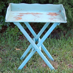 Op de afbeelding: Een lichtblauwe houten klaptafel met een verweerde afwerking. De tafel heeft een rechthoekig blad en een eenvoudige, X-vormige basis.