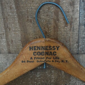 Puede incluir: Percha de ropa de madera con un acabado marr&oacute;n. La percha tiene un gancho de metal y el texto "Hennessy Cognac A Friend For Life 84 Proof Schieffelin & Co., N. Y." impreso en la parte superior.