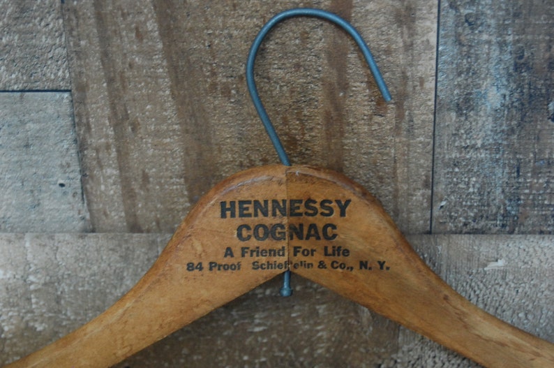 Puede incluir: Percha de ropa de madera con un acabado marr&oacute;n. La percha tiene un gancho de metal y el texto "Hennessy Cognac A Friend For Life 84 Proof Schieffelin & Co., N. Y." impreso en la parte superior.