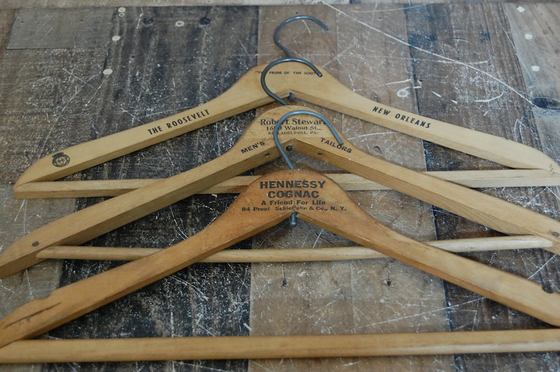 Puede incluir: Tres perchas de madera con diferentes textos publicitarios. La percha superior dice "New Orleans", la percha del medio dice "Hennessy Cognac, A Friend For Life, 84 Proof, Schieffelin & Co., N.Y.", y la percha inferior dice "The Roosevelt, Men's Tailors".