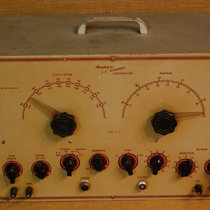 May include: A Heathkit T.W. Alignment Generator with a white face and red and black markings. The front panel has two dials, knobs, and switches. The dials are labeled "Oscillator" and "Marker". The knobs are labeled "Sweep Width", "Stand By", "Range", "Blanking", "X10", "X100", "Amplitude", "Marker", and "Range". The switches are labeled "A.C. Off", "Power", and "Output".