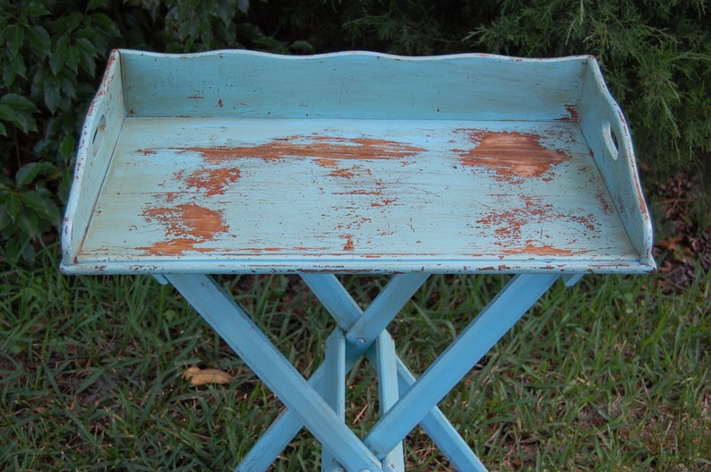 Op de afbeelding: Een blauwe houten opklapbare dienbladtafel met een verweerde afwerking. De tafel heeft een rechthoekig blad en een kruisvormig onderstel.