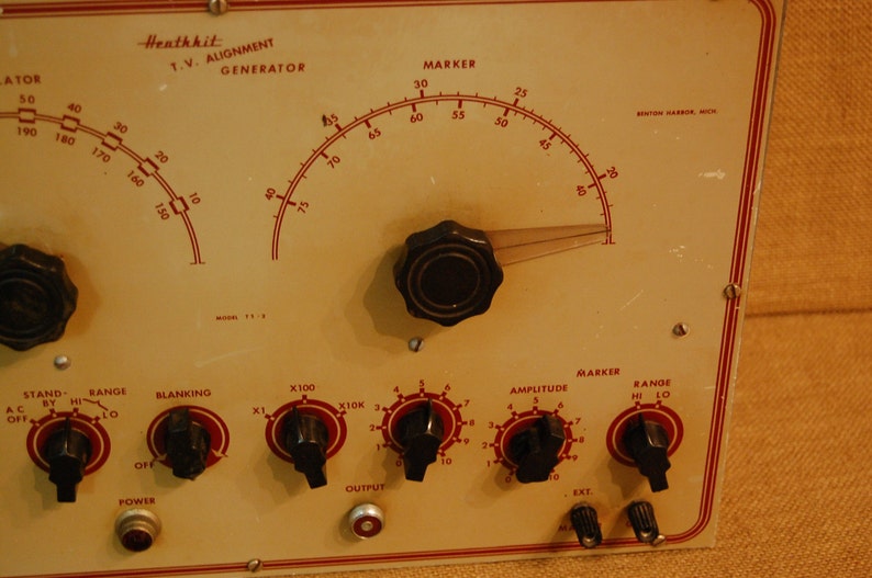 May include: A vintage Heathkit T.V. Alignment Generator with a large dial and multiple knobs. The dial is labeled "Marker" and has numbers from 25 to 75. The knobs are labeled "Stand", "Range", "Blanking", "X100", "X10K", "Amplitude", and "Marker".