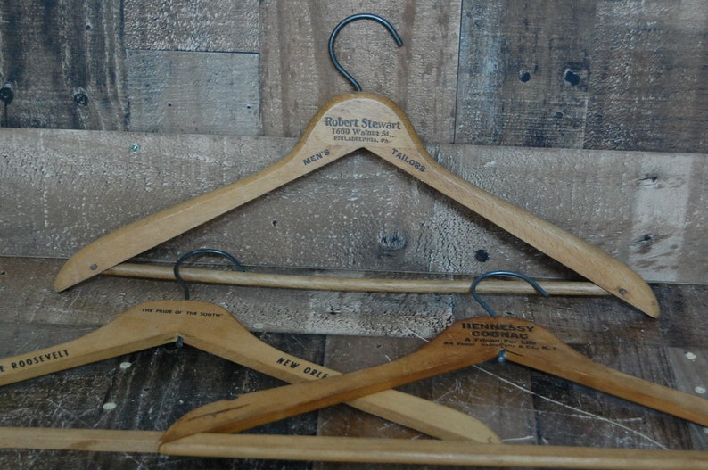 Puede incluir: Tres perchas de madera con diferentes textos publicitarios. La percha superior tiene el texto "Robert Stewart 1009 Walnut St. Philadelphia, Pa. Men's Tailors". La percha del medio tiene el texto "The Pride of the South New Orleans". La percha inferior tiene el texto "Hennessy Cognac A Friend For Life 84 Proof Produced and Bottled by Jas Hennessy & Co. Cognac, France".