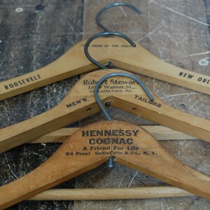Puede incluir: Tres perchas de madera con ganchos de metal. La percha superior tiene el texto "THE ROOSEVELT" y "NEW ORLEANS". La percha del medio tiene el texto "Robert L. Stewart, 160 Walnut St., Philadelphia, PA", "MEN'S" y "TAILORS". La percha inferior tiene el texto "HENNESSY COGNAC, A Friend For Life, 84 Proof Schieffelin & Co., N. Y."