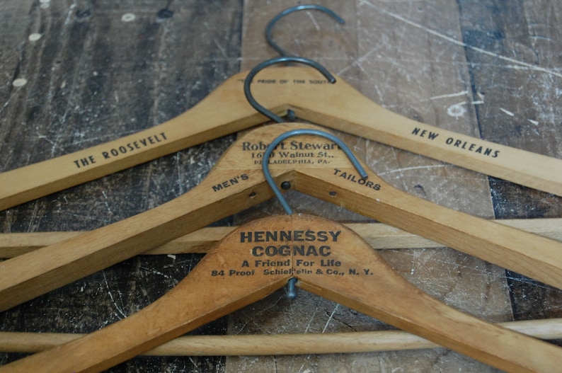 Puede incluir: Tres perchas de madera con ganchos de metal. La percha superior tiene el texto "THE ROOSEVELT" y "NEW ORLEANS". La percha del medio tiene el texto "Robert L. Stewart, 160 Walnut St., Philadelphia, PA", "MEN'S" y "TAILORS". La percha inferior tiene el texto "HENNESSY COGNAC, A Friend For Life, 84 Proof Schieffelin & Co., N. Y."