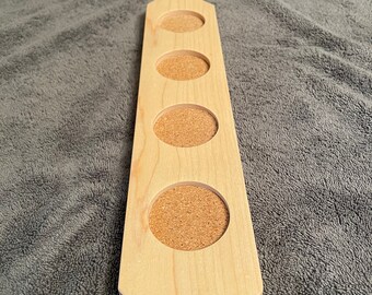 May include: A wooden paddle with four circular cutouts, each filled with a cork coaster. The paddle has a handle at the top and is made of light wood.