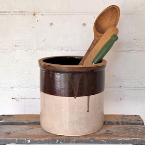 Pode incluir: Um pote de cerâmica cilíndrico com um design de dois tons, apresentando uma seção superior esmaltada em marrom escuro e uma seção inferior em bege claro. Dentro do pote, duas colheres de madeira, uma com cabo verde, estão apoiadas.