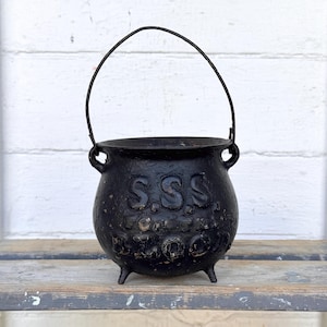 May include: A black cast iron cauldron with a curved handle and three small legs. The cauldron has the embossed letters "S.S.S." on its side. The cauldron is sitting on a weathered wooden surface against a white background.