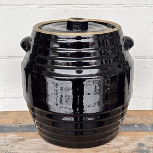 May include: A black ceramic crock with a lid. The crock has a glossy finish and a ribbed design around the body. It features two small handles and a lid with a small knob. The crock is likely used for food storage or fermentation.