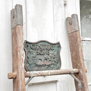 May include: Weathered wooden ladder with metal hardware, leaning against a white door. A bronze-colored sign reading "CENTURY HOME" is attached to the ladder, featuring a detailed landscape scene. The ladder's aged appearance suggests vintage or antique origins.