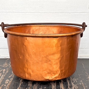 May include: A large, antique copper cauldron with a hammered finish. The pot has a rounded body and a sturdy, dark metal handle. The copper has a warm, orange-brown hue, and the cauldron sits on a dark, weathered wooden surface.